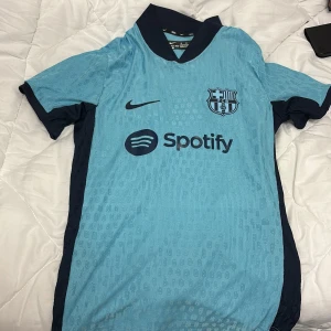 Blå FC Barcelona Nike fotbollströja S - Snygg FC Barcelona fotbollströja från Nike i ljusblått med mörkblå detaljer och krage. Tröjan har korta ärmar, klubbmärke på bröstet och Spotify-logga framtill. Tillverkad i lätt och ventilerande material, perfekt för match eller träning.