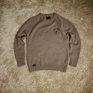 Beige stickad tröja från Morris - Snygg beige stickad tröja från Morris med rund halsringning och ribbade muddar. Tröjan har ett broderat märke på bröstet med brittisk flagga och text. Perfekt för dig som gillar klassisk och stilren look.