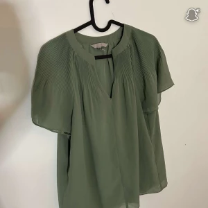 Grön blus från H&M med v-ringning - Säljer en luftig grön blus från H&M i storlek S. Blusen har korta ärmar, v-ringning och fina plisserade detaljer upptill. Tillverkad i ett lätt och flowy material som ger en chill vibe. Perfekt att styla med jeans eller kjol.