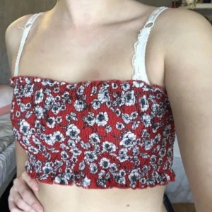 Röd blommig bandeau tubtopp - Supersöt röd bandeau tubtopp med vita och svarta blommor. Smockad och elastisk för en tight passform, perfekt till sommaren. Toppens blommiga mönster ger en lekfull vibe och den är ärmlös för en luftig känsla.