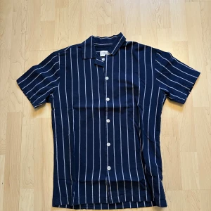 Randig blå skjorta från Lindbergh - Snygg kortärmad skjorta från Lindbergh i marinblått med vita vertikala ränder. Skjortan har klassisk krage och knäppning framtill med vita knappar. Perfekt för en avslappnad och stilren look.