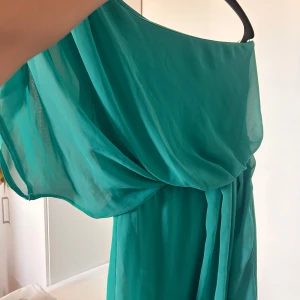 Turkos oneshoulder långklänning - Säljer en turkos kort klänning med oneshoulder-design och draperad överdel. Klänningen har ett luftigt och svepande tyg som ger en elegant siluett. Perfekt för dig som vill sticka ut med en cool färg och unik axelmodell.