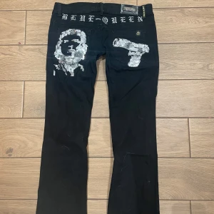 Svarta Blue Queen jeans med paljettdetaljer - Svarta jeans från Blue Queen med unika paljettdetaljer på bakfickorna – ena sidan har ett ansikte och andra en pistol i silverpaljetter. Snygg brodyr med texten 'BLUE QUEEN' baktill i vitt. Klassisk femficksmodell med raka ben och låg midja.
