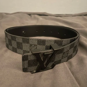 L V bälte - Snyggt L V bälte i svart och grått med klassiskt rutigt mönster och stor L V-logga i metall som spänne. Bältet är gjort i  skinn och har en modern, stilren vibe. Perfekt accessoar för att lyfta din outfit med en lyxig touch.