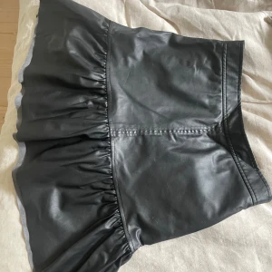 Svart volangkjol i skinnimitation H&M - Supersnygg svart kortkjol från H&M i skinnimitation med volangkant nertill. Kjolen har en markerad midja och stängs med tryckknappar i sidan. Perfekt för dig som vill ha en edgy och trendig look.