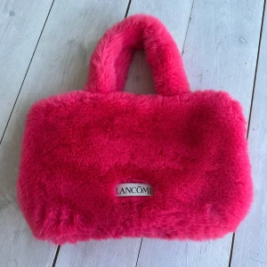 Oanvänd rosa fluffig handväska från Lancôme - Supermjuk och fluffig rosa handväska från Lancôme med pälsimitation och guldig dragkedja. Väskan har ett innerfack med dragkedja och ett Lancôme-märke framtill. Så fin! Oanvänd, går inte att köpa 