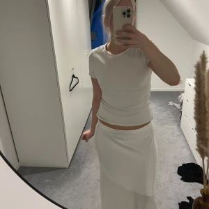 Vit långkjol i satin-look - Supersnygg vit långkjol med slät och glansig satin-feeling. Kjolen har en enkel och clean design med lätt omlott-effekt framtill. Perfekt för dig som gillar minimalistisk och stilren vibe.