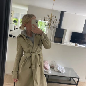 Beige trenchcoat med bälte och knappar - Stilren beige trenchcoat med klassisk krage, dubbelknäppning och bälte i midjan. Jackan har långa ärmar med spännen vid handlederna och slits bak. 