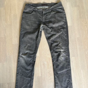 Nudie Jeans Thin Finn grå jeans W32 L32 - Slimmade Thin Finn jeans från Nudie Jeans i en snygg grå tvätt med coated finish. Klassisk femficksmodell, normal midja och raka ben. Materialet är 98% bomull och 2% elastan för lite stretch. Perfekta för dig som gillar en clean och modern look. Hål i skrevet som är lappat men fortfarande synligt.