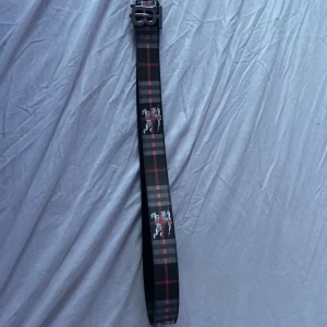 Rutig bälte med metallspänne - Säljer ett snyggt rutig Burberrybälte med ett coolt metallspänne. Bältet har ett klassiskt rutmönster i svart, grått och rött, perfekt för att ge din outfit en extra edge. Det är i bra skick och passar perfekt till både vardag och fest. Spännet har en unik design som verkligen sticker ut! Bältet passar ungefär 10-14 åringar. Priset kan diskuteras vid snabb affär.