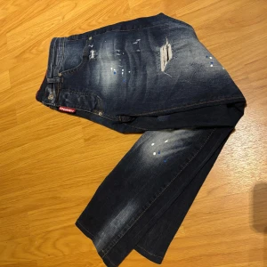 Dsquared2 blå distressed jeans - Snygga blå jeans från Dsquared2 med slitna detaljer och färgstänk i vitt och blått. Jeansen har en smal passform, klassisk femficksdesign och coola slitningar på benen. Perfekta för dig som gillar en edgy streetstil. (STORLEK 30/31)