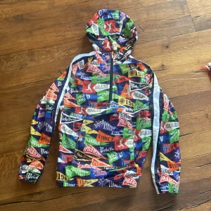 Färgglad vindjacka Polo Ralph Lauren - Säljer en riktigt färgstark vindjacka från Polo Ralph Lauren med huva och halv dragkedja. Jackan har ett unikt collage-mönster med olika loggor och text i blått, grönt, rött, gult och orange. Tillverkad i polyester och har vita ränder längs ärmarna.