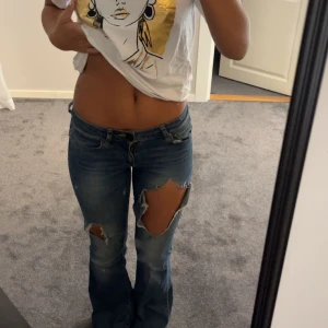 Blå bootcut jeans med stora hål - Säljer ett par blå bootcut jeans med slitna och stora hål på låret och knät. Jeansen har låg midja, klassisk knapp och dragkedja framtill samt fem fickor.  Byxan har gått sönder i bak. Det finns ett litet hål i byxan där skärp grejen ska sitta. 