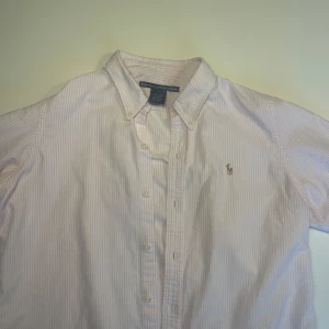 Rosa randig skjorta från Ralph Lauren - Klassisk rosa och vit randig skjorta från Ralph Lauren med button-down krage och broderad logga på bröstet. Skjortan har långa ärmar och knäppning framtill. Perfekt för en clean och stilren look.