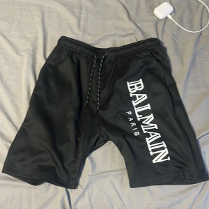 Svarta shorts från Balmain Paris - Snygga svarta shorts från Balmain Paris med stor vit logga längs benet. De har elastisk midja med snörning och är tillverkade i ett mjukt, glansigt syntetmaterial. Perfekta för dig som vill ha en clean och lyxig streetwear-look.