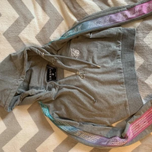 Grå hoodie från SikSilk med färgglada detaljer - Säljer en grå hoodie från SikSilk med dragkedja och snörning i huvan. Tröjan har långa ärmar med coola färgglada band i lila och turkos längs sidorna där det står SikSilk. Mjuk bomullskänsla och broderad logga på bröstet. Perfekt för chill eller streetwear.
