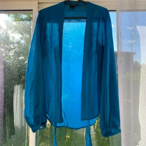 Blå transparent skjorta med knytband - Snygg blå skjorta i transparent material med långa ärmar och knytband nedtill. Skjortan har knappar framtill och manschetter med knappdetalj. Perfekt för dig som vill ha en luftig och trendig look.