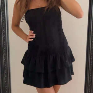 Svart axelbandslös volangklänning - Snygg svart kortklänning med axelbandslös design och dubbla volanger nertill. Klänningen har en smockad överdel som ger en tight passform och en feminin siluett. Perfekt för dig som vill ha en enkel men trendig look.