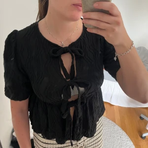 Svart blus med knyt och struktur - Säljer en svart blus med korta puffärmar och flera knyt framtill. Tyget är strukturerat och ger en cool, vågig look. Perfekt för dig som gillar unika detaljer och en trendig stil.