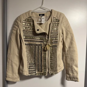 Beige jacka med paljettdetaljer Lindex - Snygg beige jacka från Lindex med unika paljettdetaljer och mönstrade dekorationer framtill. Jackan har dragkedja med tofs och quiltade axlar för en cool look. Perfekt för dig som vill sticka ut med stilrena detaljer och trendig design.