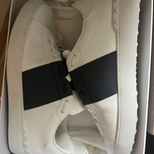 Valentino Garavani Open  - Säljer ett par snygga Valentino Garavani Open sneakers i vitt skinn med bred svart rem över mitten. vita snören och diskreta nitar på sulan. Kommer med originalkartong och dustbag. Skorna passar till de mesta och är super snygg