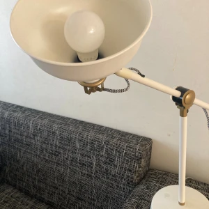 Justerbar Bordslampa, metall - Stilren vit och mässingsfärgad skrivbordslampa med justerbar arm och skärm. Passar perfekt för arbetsplats eller läshörna. Lampan är i gott skick med mindre spår av användning. Levereras med LED-lampa. Snygg retrodesign och stabil fot. Nypris 699 kr 