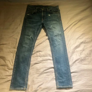 Blå skinny jeans från Nudie Jeans - Säljer ett par blå skinny jeans från Nudie Jeans med slitna detaljer och klassisk femficksdesign. Jeansen är tillverkade i 99% bomull och 1% elastan för en bekväm stretch. Snygg tvätt och smal passform som sitter tajt längs benen.