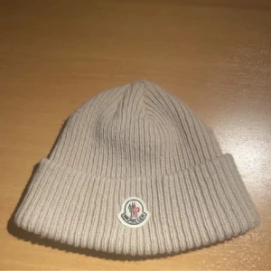 Beige Moncler mössa i ull - Snygg beige mössa från Moncler med ribbad stickning och uppvikt kant. Klassisk Moncler-logga framtill och diskret etikett på insidan. Perfekt för dig som vill ha en clean och lyxig look under kalla dagar.