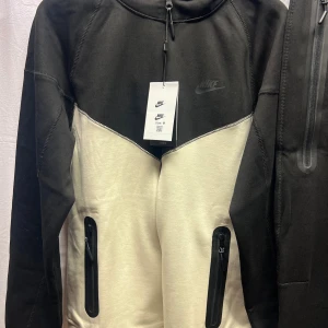 Nike hoodie i svart och beige - Nike hoodie i svart och beige med dragkedjor på fickorna och snygg tvåfärgad design. Tröjan har huva, långa ärmar och sportig look. Perfekt för dig som gillar streetwear och vill ha något bekvämt och stilrent.