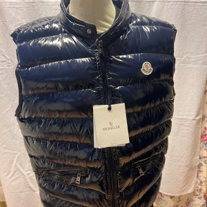 Mörkblå dunväst från Moncler - Snygg mörkblå dunväst från Moncler med glansig finish och klassisk logga på bröstet. Västen har ståkrage, två dragkedjeförsedda fickor och quiltad design. Perfekt för lager-på-lager och har en cool, sportig vibe. Dragkedja framtill och innerficka med serietidningsetikett.