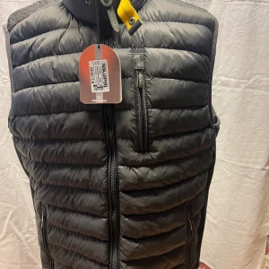 Svart dunväst från Parajumpers XL - Svart dunväst från Parajumpers i storlek XL med horisontella quiltade paneler och gul detalj vid kragen. Västen har flera dragkedjeförsedda fickor och en snygg logotyp på nacken. Perfekt för lager-på-lager och chill stil.
