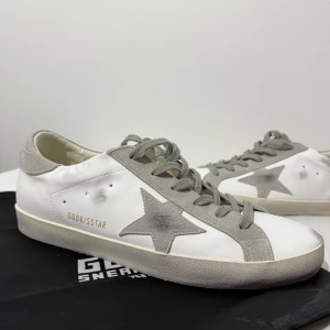 Golden Goose Superstar sneakers vit/grå - Golden Goose Superstar sneakers i vitt skinn med grå mockadetaljer och snörning. Ikonisk grå stjärna på sidan och textdetaljer i guld. Platt sula och rund tå. Snyggt slitna detaljer för en cool look. Perfekt för dig som gillar italiensk design och streetstyle. Säljer dem pga av att dem är stora på mig. Passar som 43 också. Aldrig använt, helt nya.  