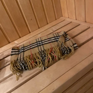 Beige rutig halsduk med fransar - Snygg beige halsduk från Burberry med klassiskt rutigt mönster i svart, vitt och rött. Mjuk och varm, perfekt att slänga runt halsen när det är kallt ute. Fransar i ändarna ger en chill vibe. Passar dig som gillar tidlös stil och vill ha något som sticker ut.