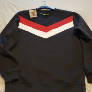 Svart sweatshirt med röd och vit detalj - Svart sweatshirt från Armygross i storlek L med en snygg V-formad röd och vit panel över bröstet. Tröjan har ribbade muddar vid hals, ärmslut och nederkant. Insidan är mjuk och fleecefodrad för extra komfort. Perfekt för dig som gillar stilrena och sportiga vibes.