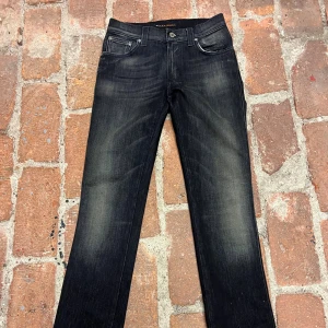Svarta jeans från Nudie Jeans Co, 28/32 - Snygga svarta jeans från Nudie Jeans Co i storlek 28/32. Jeansen har en klassisk femficksdesign, raka ben och diskreta slitningar framtill. Bakfickorna har Nudie Jeans signatursöm. Perfekta för dig som gillar stilrena och tidlösa jeans. Alldrig använda!