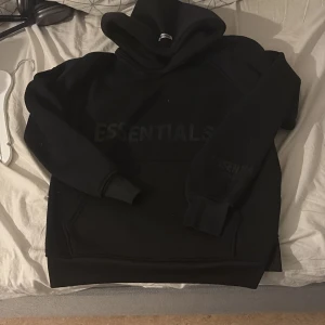 Svart Essentials hoodie med huva - Svart hoodie från Essentials med stor huva och tryckt logga på bröstet. Tröjan har en klassisk känguruficka framtill och ribbade muddar vid ärmslut och nederkant. Perfekt för chill dagar och streetwear-stil.