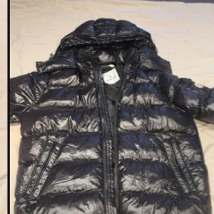 Svart pufferjacka från Moncler - Säljer en svart, glansig pufferjacka från Moncler med huva och dragkedja. Jackan har Moncler-logga på ärmen och coola detaljer som serietidningsmönster på insidan. Perfekt för dig som vill ha en varm och snygg vinterjacka med streetkänsla.