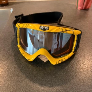 Gula Zipe skidglasögon barn  - Säljer ett par coola goggles från Zipe Airways med gul ram och svarta ränder. Linsen är spegeltonad med orange insida och har bred svart elastisk rem med logotyp. Perfekt för dig som vill sticka ut i backen eller på crossbanan.