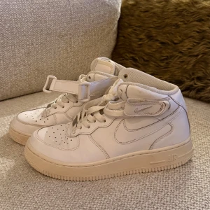 Nike Air Force 1 Mid  - Nike Air Force 1 Mid sneakers i helvitt skinn med klassisk snörning och kardborreband runt ankeln. Snygg, chunky siluett med perforerad tå och platt sula. Ikonisk Nike Swoosh på sidan och AIR-logga bak. Perfekt för dig som gillar streetstyle.