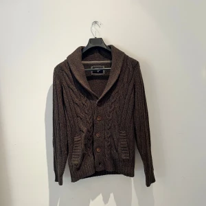 Brun stickad kofta från Cedar Wood State - Säljer en brun stickad kofta från Cedar Wood State i storlek M. Koftan har grovt kabelstickat mönster, bred krage och tre knappar framtill. Två fickor på sidorna och långa ärmar ger en chill och klassisk look. Perfekt för lager-på-lager under kyliga dagar. Hör av er vid fler frågor eller fler bilder
