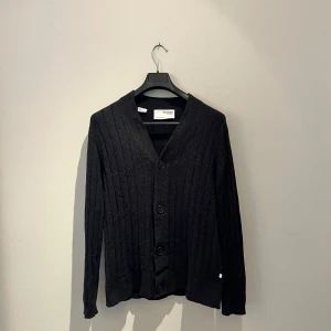 Svart ribbad kofta från Selected Homme - Svart kofta från Selected Homme i storlek L. Tröjan är ribbstickad med vertikala ränder, har v-ringning och stora svarta knappar framtill. Långärmad och tillverkad i mjukt stickat material som känns skönt mot huden. Perfekt för dig som gillar stilrena och enkla plagg. Hör av er vid fler bilder eller frågor