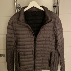 Grå pufferjacka med dragkedja - Säljer en stilren grå pufferjacka med horisontella sömmar och svarta dragkedjor på fickorna. Jackan har en hög krage och normal passform, perfekt för kyliga dagar. Enkel och snygg design som funkar till det mesta.