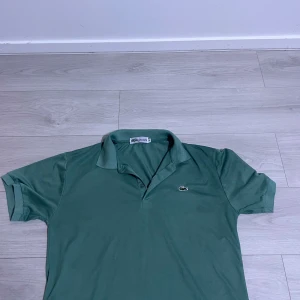 Grön pikétröja från Lacoste - Snygg grön pikétröja från Lacoste med klassisk krage och korta ärmar. Tröjan har det ikoniska krokodilmärket broderat på bröstet och är tillverkad i bomull för en skön och luftig känsla. Perfekt för en stilren och avslappnad look.