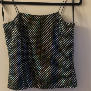 Glittrig svart prickig linnetopp - Säljer en svart linnetopp med tunna axelband och glittriga prickar över hela tyget. Prickarna har en holografisk effekt som reflekterar regnbågens färger. Toppen är rak i modellen och har en cool partyvibe. Perfekt för dig som vill sticka ut med lite extra bling.