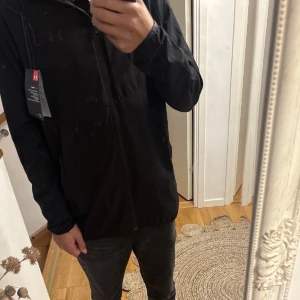 Svart vindjacka från Under Armour - Svart vindjacka från Under Armour med huva och hel dragkedja framtill. Jackan har diskret logga på bröstet och tryckt Under Armour-text på ryggen. Tillverkad i lätt syntetmaterial, perfekt för blåsiga dagar. Justerbar huva med snörning och två sidofickor.