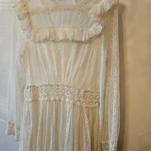 Vit spetsklänning från By Malina - Superfin vit spetsklänning från By Malina med romantiska volangdetaljer, markerad midja med broderade blommor och långa ärmar. Klänningen har en kort längd och är helfodrad med transparent spets över hela. Perfekt för dig som gillar en feminin och bohemisk stil.