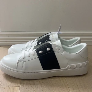 Valentino Garavani Open sneakers vit/svarta - Snygga Valentino Garavani Open sneakers i vitt skinn med bred marinblå rem över vristen och ikoniska nitar på hälen. Klassisk rund tå, vita snören och guldlogga på plösen. Perfekta för dig som vill ha en clean men lyxig look.