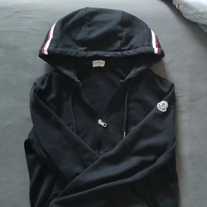 Marinblå hoodie från Moncler med dragkedja - Svart hoodie från Moncler med vit och vinröd rand på huvan. Klassisk logga på ärmen och dragkedja framtill. Skön passform och perfekt för dig som gillar stilrena streetwear-plagg. Tillverkad i mjukt bomullsmaterial.