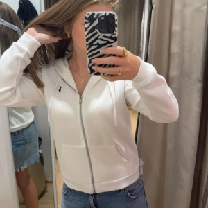 Vit hoodie från Ralph Lauren - Säljer en fräsch vit hoodie från Ralph Lauren med dragkedja, huva och snörning. Tröjan har två fickor framtill och den klassiska blå Polo-loggan på bröstet. Perfekt till jeans och sneakers för en chill look.