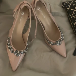 Pumps med strass från Dancini - Snygga ljusrosa pumps från Dancini med spetsig tå och dekorativa strassdetaljer framtill. Skorna har en elegant klack och smala remmar runt ankeln som ger en chic look. Perfekta för dig som gillar att sticka ut med glittriga detaljer.
